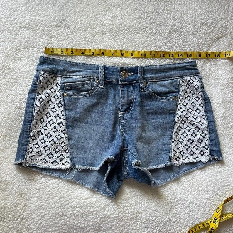 May include: Light blue denim shorts with white lace panels on the sides. The shorts have a raw hem and classic five-pocket styling. The shorts measure approximately 18 inches across.