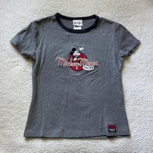 May include: A heather grey short-sleeve t-shirt with a black crew neckline. The chest features an embroidered Mickey Mouse graphic over a red circle with the text Mickey Mouse Since 1928. A small rectangular Authentic Mickey label is at the hem.