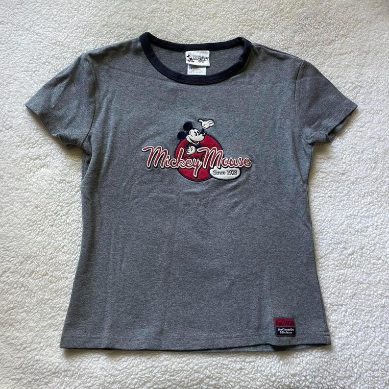 May include: A heather grey short-sleeve t-shirt with a black crew neckline. The chest features an embroidered Mickey Mouse graphic over a red circle with the text Mickey Mouse Since 1928. A small rectangular Authentic Mickey label is at the hem.