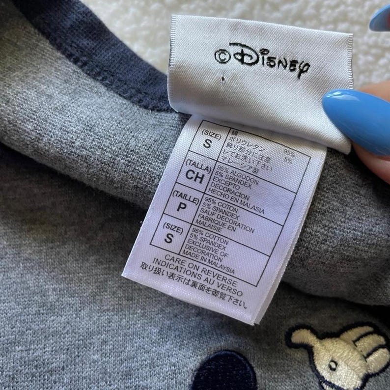 May include: A grey heathered garment with a dark grey trim and a white Disney brand tag. The tag displays size S and fabric composition of 95% cotton and 5% spandex, with care instructions in multiple languages including English, French, and Japanese.