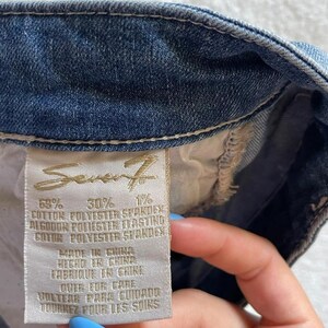 May include: Close-up of a blue denim garment with a tag. The tag features the brand name "Seven7" and fabric composition details. The tag also includes instructions in multiple languages: English, Spanish, and French.