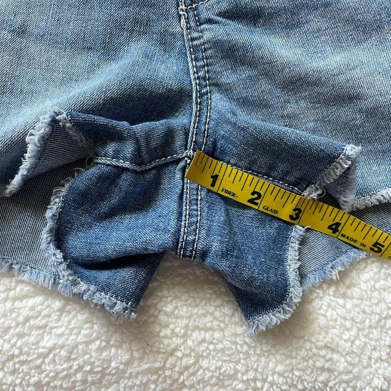 May include: Close-up of light blue denim shorts with frayed edges. A yellow measuring tape shows a measurement of approximately 3 inches. The shorts have white stitching and are on a white, fuzzy surface.