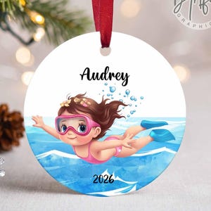 May include: A round white ceramic ornament hanging from a red ribbon. The design features a cartoon child swimming in blue water, wearing pink goggles and a pink swimsuit. The text Audrey is printed above the child, and 2026 is below.
