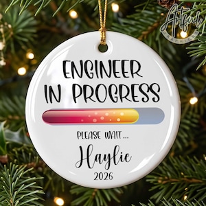May include: A round white ceramic ornament featuring the black text ENGINEER IN PROGRESS above a colorful loading bar with a gradient from red to yellow. Below the bar, the text reads PLEASE WAIT... followed by the name Haylie and the year 2026.