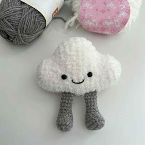Puede incluir: Un peluche hecho a mano con forma de nube blanca con ojos negros y una boca sonriente. La nube tiene patas de ganchillo grises. Dos ovillos de lana, uno gris y otro blanco, están en el fondo.