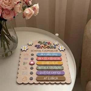 May include: A wooden status sign for a school counselor named Mrs. Brooks, featuring a scalloped edge, white daisy accents, and six colorful horizontal bars with text options including In a Meeting, In a Classroom, Come on In, With a Student, Out of Office, and At Lunch.
