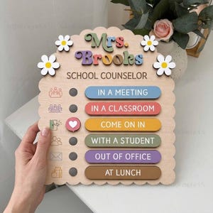 May include: A light-colored wooden sign with scalloped edges featuring the text Mrs. Brooks and School Counselor. It includes four white flower cutouts and six horizontal status bars with icons, including In a Meeting, In a Classroom, Come on In, With a Student, Out of Office, and At Lunch.