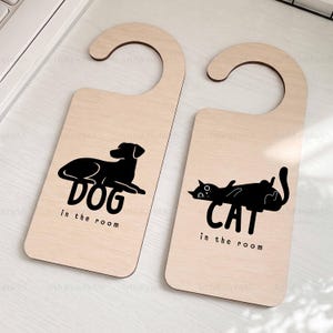May include: Two light-colored wooden door hangers with black graphic designs. One features a silhouette of a dog with the text DOG in the room, and the other features a silhouette of a cat with the text CAT in the room, both with rounded hooks.