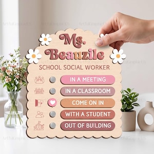 May include: A scalloped wooden sign featuring the text Ms. Beauzile SCHOOL SOCIAL WORKER and five status options: IN A MEETING, IN A CLASSROOM, COME ON IN, WITH A STUDENT, and OUT OF BUILDING, with a heart icon indicating the current status.