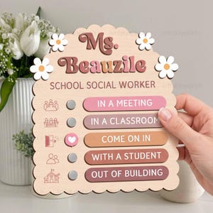May include: A light-colored wooden sign with scalloped edges featuring the text Ms. Beauzile School Social Worker. It includes five status options with circular markers, one of which is selected with a heart icon, and white flower accents.