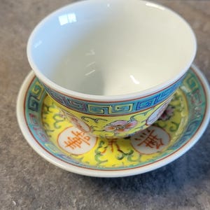 May include: A white teacup and saucer set with a yellow and teal floral design. The teacup sits on a matching saucer with Chinese characters. The set is likely porcelain.