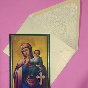 Può includere: Un'icona della Vergine Maria e del bambino, incorniciata in verde e oro, con una croce. L'icona è accompagnata da una busta color crema con un motivo a croci. Lo sfondo è rosa.