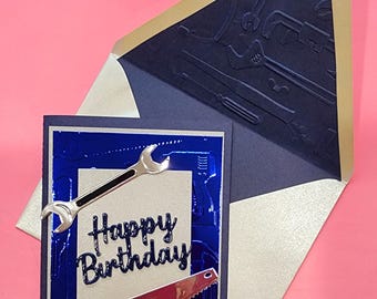 Op de afbeelding: Een blauwe kaart met een zilveren frame en een zilveren achtergrond. De kaart heeft een moersleutel en een zaag erop. De tekst "Happy Birthday" staat in zilveren letters op de kaart.