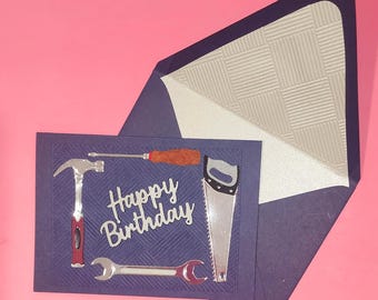 Op de afbeelding: Een donkerblauwe kaart met een zilveren reliëfpatroon op de envelop. De kaart heeft een zilveren en rode hamer, schroevendraaier, moersleutel en zaag met de tekst "Happy Birthday" in zilveren letters.