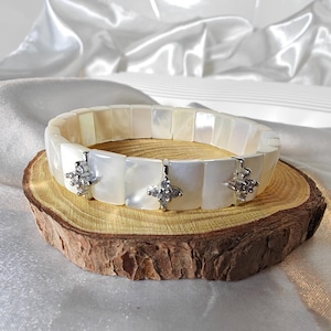 May include: A white mother-of-pearl bracelet with square segments and silver-colored, four-petal flower accents. The bracelet is displayed on a wooden slice against a white satin background.