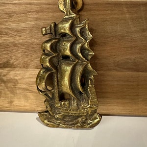 May include: A gold-toned brass door knocker shaped like a sailing ship with intricate details. The ship has multiple sails and a detailed hull.