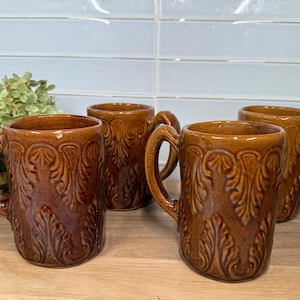 May include: Four brown ceramic mugs with ornate leaf and scroll designs. Each mug has a large handle and a glossy finish. The mugs are arranged on a wooden surface, with a light blue tile background.