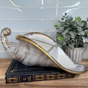 May include: A decorative, horn-shaped ceramic vase with a white and gold finish. The vase is placed on a dark blue book with gold accents. A small potted plant is in the background.