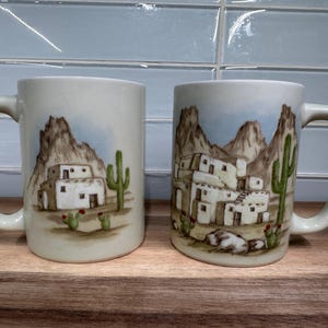 May include: Two ceramic mugs with a Southwestern desert scene. Each mug features a landscape with white buildings, cacti, and mountains in shades of brown, green, and blue. The mugs have a cream-colored background and large handles.
