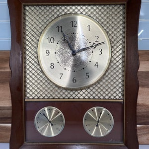 May include: A rectangular wooden clock with a gold-colored face and a decorative mesh background. The clock face has black numerals and hands, with two smaller dials below. The frame is a dark brown color.
