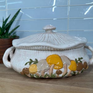 May include: White ceramic casserole dish with a lid featuring a mushroom design. The mushrooms are painted in shades of brown, yellow, and white.