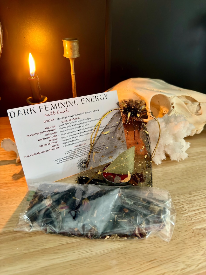 K&ouml;nnte beinhalten: Ein Dark Feminine Energy Salzschalen-Set bestehend aus einem schwarzen Organza-Beutel mit goldenen Sternenmustern, einem durchsichtigen Plastikbeutel mit getrockneten Kr&auml;utern und Bl&uuml;tenbl&auml;ttern, einer roten Kerze, einem Lorbeerblatt und einer Infokarte.