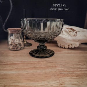 K&ouml;nnte beinhalten: Eine rauchgraue Glasschale mit einem Standfu&szlig;, die ein strukturiertes Muster aus vertikalen Rillen und kreisf&ouml;rmigen Motiven aufweist. Der Text "STYLE C: smoke gray bowl" ist in Wei&szlig; &uuml;ber der Schale aus durchscheinendem Glas gedruckt.