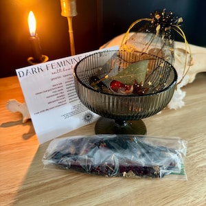 K&ouml;nnte beinhalten: Eine dunkelgr&uuml;ne, gerippte Glasschale auf einem Fu&szlig;, die ein Lorbeerblatt und ein kleines rotes Objekt enth&auml;lt, begleitet von einem schwarzen Organza-Beutel mit goldenen Sternenmustern, einer wei&szlig;en Infokarte mit der Aufschrift DARK FEMININE und Kr&auml;utern.