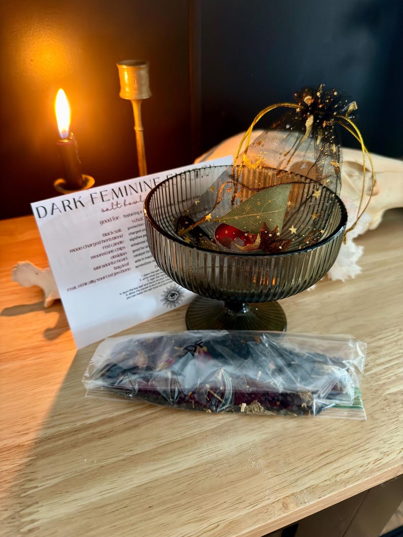 K&ouml;nnte beinhalten: Eine dunkelgr&uuml;ne, gerippte Glasschale auf einem Fu&szlig;, die ein Lorbeerblatt und ein kleines rotes Objekt enth&auml;lt, begleitet von einem schwarzen Organza-Beutel mit goldenen Sternenmustern, einer wei&szlig;en Infokarte mit der Aufschrift DARK FEMININE und Kr&auml;utern.