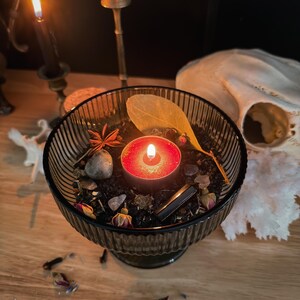 K&ouml;nnte beinhalten: Eine gerippte Schale aus dunklem Glas, gef&uuml;llt mit schwarzem Wachs, einem brennenden roten Teelicht, einem getrockneten Blatt, kleinen Rosenknospen, einem Sternanis, einem glatten grauen Stein und einem dunklen Kristall, umgeben von Streublumen.