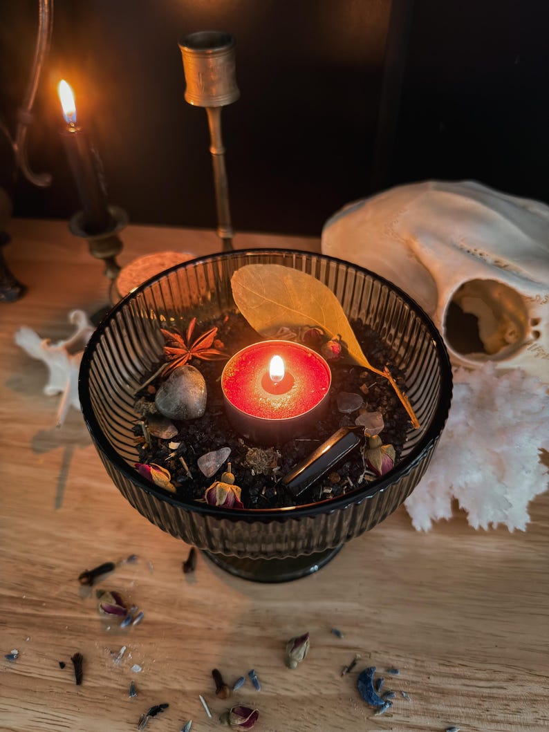 K&ouml;nnte beinhalten: Eine gerippte Schale aus dunklem Glas, gef&uuml;llt mit schwarzem Wachs, einem brennenden roten Teelicht, einem getrockneten Blatt, kleinen Rosenknospen, einem Sternanis, einem glatten grauen Stein und einem dunklen Kristall, umgeben von Streublumen.