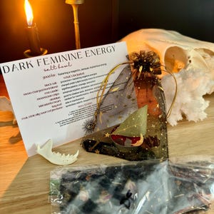 K&ouml;nnte beinhalten: Ein Set mit der Aufschrift Dark Feminine Energy, bestehend aus einer wei&szlig;en Karte mit schwarzem Text, einem schwarzen Organzabeutel mit goldenen Sternen, einer roten Kerze, Kristallen, einem Lorbeerblatt, einem Knochenst&uuml;ck und Kr&auml;utern.