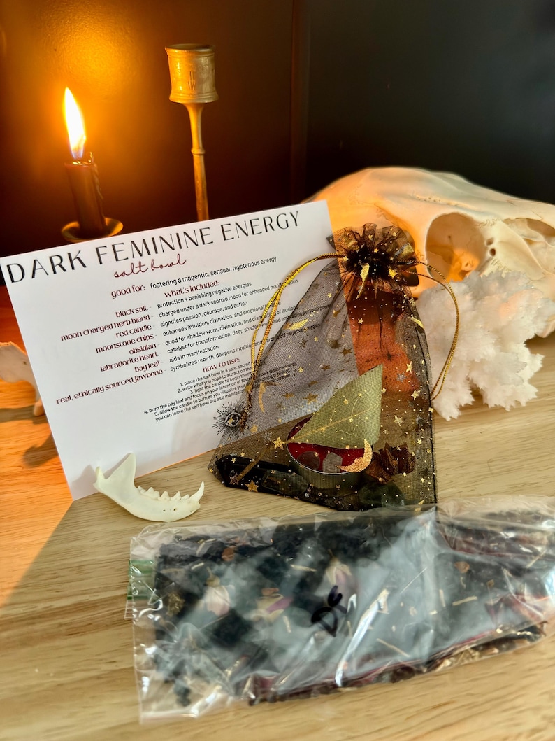 K&ouml;nnte beinhalten: Ein Set mit der Aufschrift Dark Feminine Energy, bestehend aus einer wei&szlig;en Karte mit schwarzem Text, einem schwarzen Organzabeutel mit goldenen Sternen, einer roten Kerze, Kristallen, einem Lorbeerblatt, einem Knochenst&uuml;ck und Kr&auml;utern.
