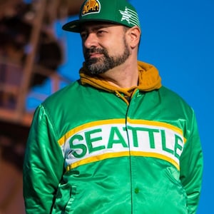 May include: A man wearing a green satin bomber jacket with the word "SEATTLE" in white letters on the front. He is also wearing a green and yellow baseball cap with a star on the front.