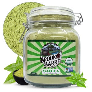 May include: A clear glass jar filled with green matcha powder, labeled "Brook & Barrel Matcha Premium Organic Tea." The jar is next to a small black bowl of matcha and fresh green tea leaves. A circle of matcha powder is in the background. The jar contains 25 oz of matcha.