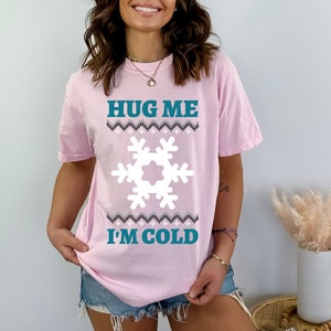 May include: A light pink short-sleeve t-shirt featuring the teal text HUG ME I'M COLD, a large white snowflake graphic, and a horizontal band with a black, white, and gray geometric pattern above and below the snowflake design.