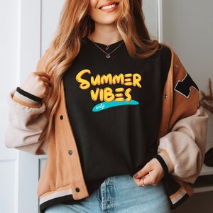 May include: A black t-shirt with a yellow and blue graphic that says "Summer Vibes Only". The t-shirt is being worn by a person with long brown hair and a brown jacket.