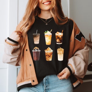 May include: A black t-shirt with a graphic of six different iced coffee drinks. The drinks are all in clear cups with different toppings and flavors. The shirt is being worn by a person with long brown hair.