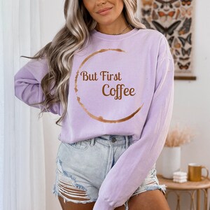 May include: A light purple sweatshirt with the phrase "But First Coffee" in brown lettering and a coffee stain design. The sweatshirt is a relaxed fit and is paired with denim shorts.