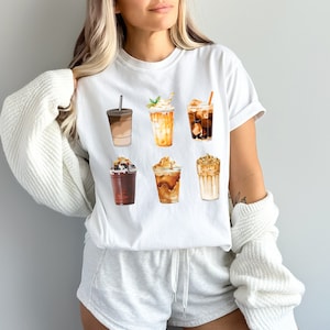 May include: White t-shirt with a graphic of six different iced coffee drinks. The drinks are illustrated in a watercolor style and include a variety of toppings, such as whipped cream, caramel, and chocolate.
