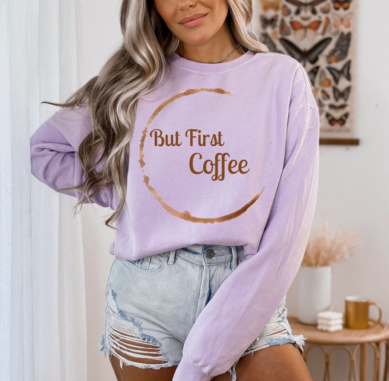 May include: A light purple sweatshirt with the phrase "But First Coffee" in brown lettering and a coffee stain design. The sweatshirt is a relaxed fit and is paired with denim shorts.