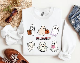May include: A white long-sleeve sweatshirt featuring a graphic print of six cartoon ghosts in various poses, including one holding a brown bag, one with an orange pumpkin bucket, and one reading a pink book. The word HALLOWEEN is printed in red.