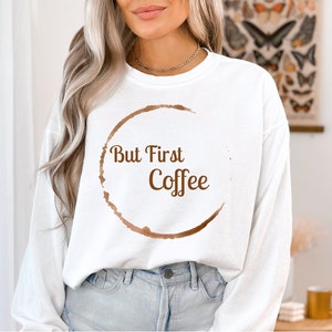 May include: White long-sleeve sweatshirt with a brown coffee stain circle design. The words "But First Coffee" are printed in brown inside the circle. The sweatshirt is a casual style.
