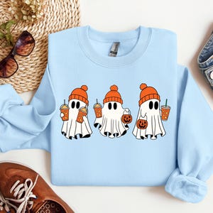 K&ouml;nnte beinhalten: Hellblauer Pullover mit drei Cartoon-Geistern, die orangefarbene H&uuml;te tragen und orangefarbene Becher und K&uuml;rbisse halten. Die Geister tragen orangefarbene H&uuml;te und halten orangefarbene Becher und K&uuml;rbisse.