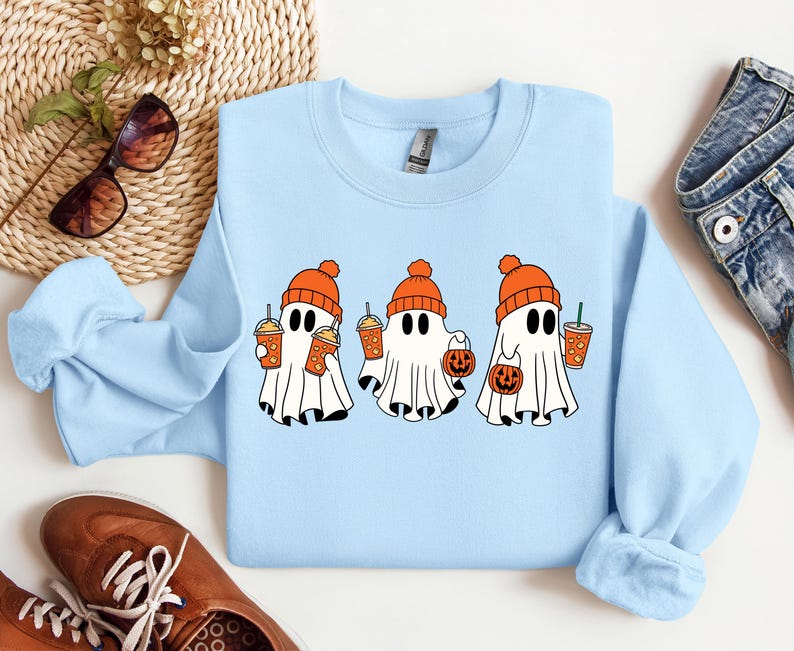 K&ouml;nnte beinhalten: Hellblauer Pullover mit drei Cartoon-Geistern, die orangefarbene H&uuml;te tragen und orangefarbene Becher und K&uuml;rbisse halten. Die Geister tragen orangefarbene H&uuml;te und halten orangefarbene Becher und K&uuml;rbisse.