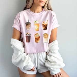 May include: Pink t-shirt with a graphic of six different iced coffee drinks. The drinks are all in different cups and have different toppings, such as whipped cream, caramel, and chocolate.