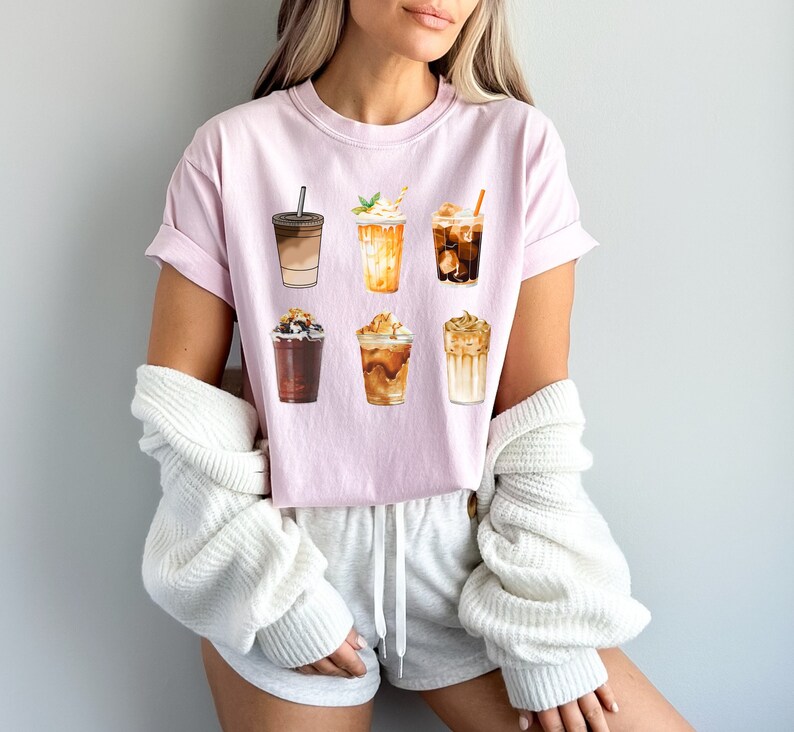 May include: Pink t-shirt with a graphic of six different iced coffee drinks. The drinks are all in different cups and have different toppings, such as whipped cream, caramel, and chocolate.