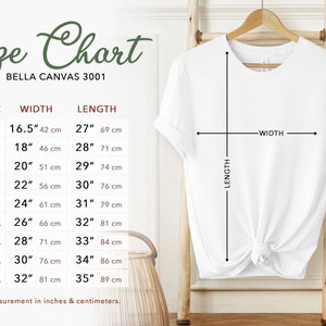 May include: A white t-shirt hanging on a wooden rack, with a size chart displayed to the left. The chart shows measurements in inches and centimeters for sizes XS to 5XL, including width and length. The text "Size Chart" is at the top.