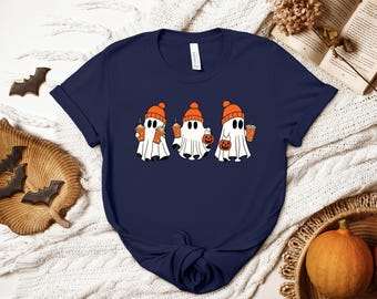 May include: A navy blue short-sleeved t-shirt featuring a graphic of three white ghosts wearing orange beanies. Each ghost is holding an orange drink cup, and one ghost is also holding a small orange jack-o'-lantern bucket for trick-or-treating.