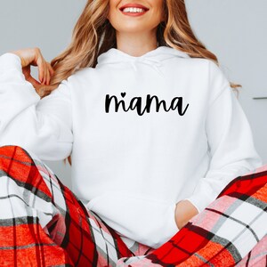 May include: White hooded sweatshirt with the word "mama" written in black cursive with a small heart above the "a". The model is wearing the sweatshirt with red and white plaid pajama pants.